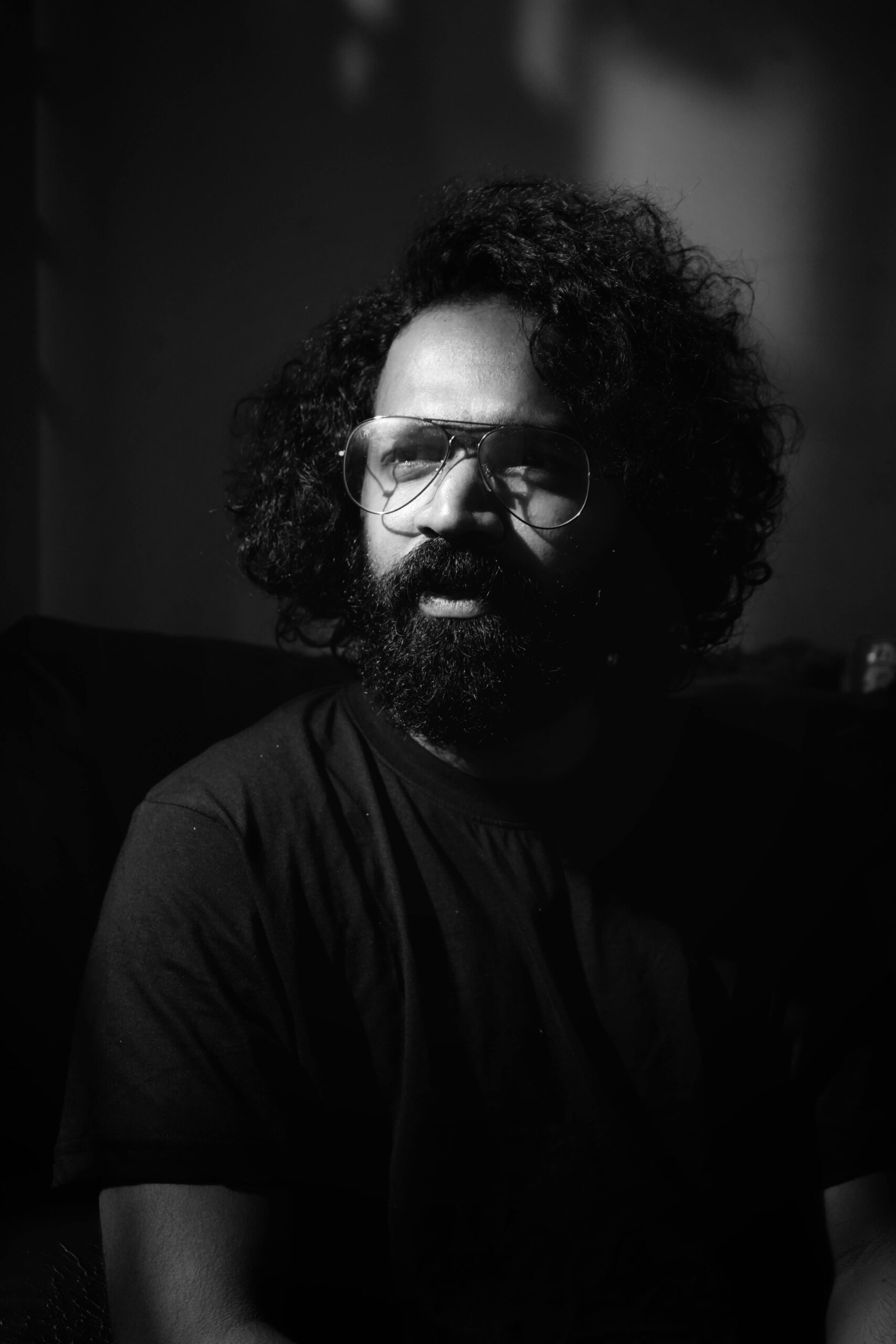 Black and white portrait of a man with curly hair and beard wearing eyeglasses. Dramatic lighting accentuates the features.
